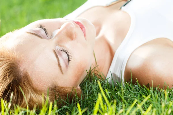 Primer plano retrato de una atractiva joven acostada sobre hierba verde en un parque durante el verano y escuchando música con sus auriculares, siendo reflexiva al aire libre. — Foto de Stock