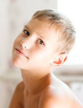 Sevimli, genç, neşeli, doğal bir gülüşü olan sarışın bir çocuk açık bej bir arka plana karşı çıplak omuzlarıyla mutlu bir şekilde gülümsüyor..