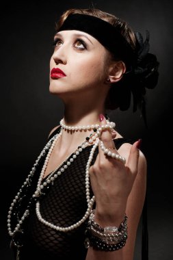 Mükemmel makyajlı, şaşaalı koyu renk saçlı bir mankenin portresi. Şık bir kolye, siyah bir kıyafet ve uzun bir bluz giyiyor. Ellerini başının arkasında tutuyor ve ileriye bakıyor..