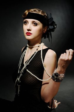 Mükemmel makyajlı, şaşaalı koyu renk saçlı bir mankenin portresi. Şık bir kolye, siyah bir kıyafet ve uzun bir bluz giyiyor. Ellerini başının arkasında tutuyor ve ileriye bakıyor..