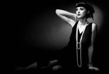 Mükemmel makyajlı, şaşaalı koyu renk saçlı bir mankenin portresi. Şık bir kolye, siyah bir kıyafet ve uzun bir bluz giyiyor. Ellerini başının arkasında tutuyor ve ileriye bakıyor..