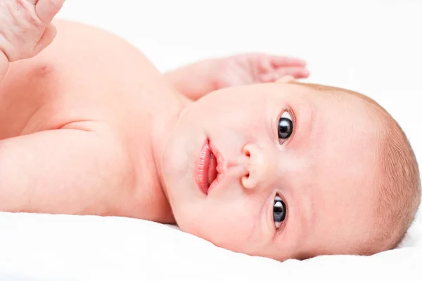 Il bambino nudo giace sulla schiena su un lenzuolo bianco e mostra emozioni. Concentrazione selettiva sulla testa dei bambini. Il bambino guarda direttamente la telecamera. Primo piano. — Foto Stock