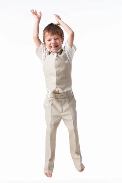 Um garotinho bonito vestido de estudante - com uma camisa branca e calças cinzas, encosta-se a uma parede branca e posa, pula como um modelo. isolado em fundo branco. — Fotografia de Stock