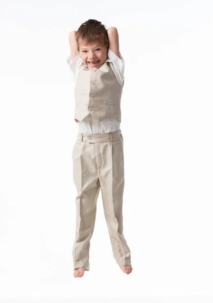 Um garotinho bonito vestido de estudante - com uma camisa branca e calças cinzas, encosta-se a uma parede branca e posa, pula como um modelo. isolado em fundo branco. — Fotografia de Stock