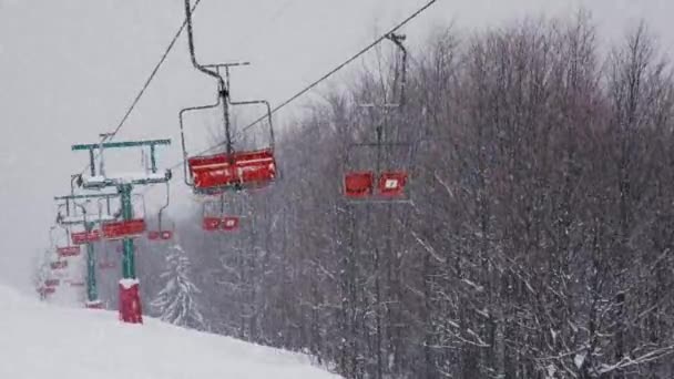 空のスキーリフトが動き雪が天から降って曇りの冬の日に下からの眺め スキー場の人がいないワーキングチェアリフトの背景に雪の雲 高品質 — ストック動画