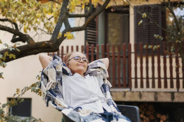 田舎の秋の庭にあるアームチェアに腰かけながらリラックスした大人の笑顔の女性 — ストック写真