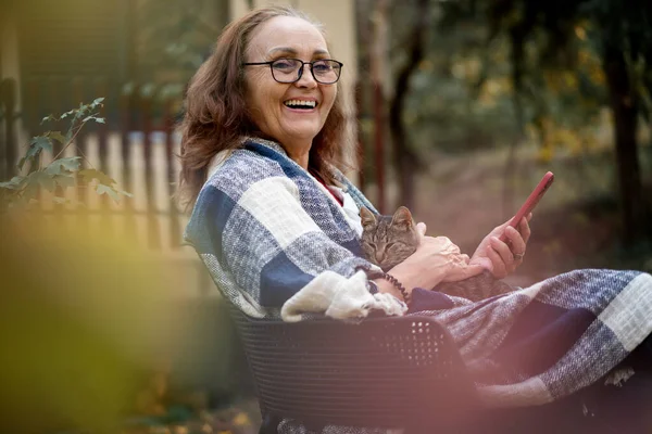Mulher sorridente feliz madura bonita com um gato em seus braços se comunicando usando um smartphone on-line no jardim de outono da casa — Fotografia de Stock