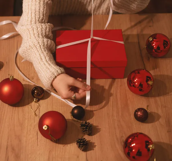 Christmas and new year gift concept, female hands with red box and white ribbon on wooden table with red christmas balls — Stock Photo, Image