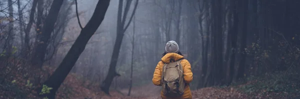 Jeune voyageuse vêtue d'une veste jaune avec sac à dos debout dans la forêt brumeuse avec le dos. Format de bannière — Photo