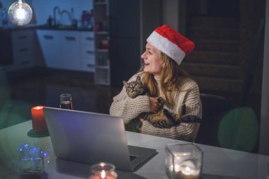 Noel Baba şapkalı kedisi olan güzel genç bir kadın Noel 'i ve yeni yılı internette kutlayan bir dizüstü bilgisayarın önünde oturuyor.