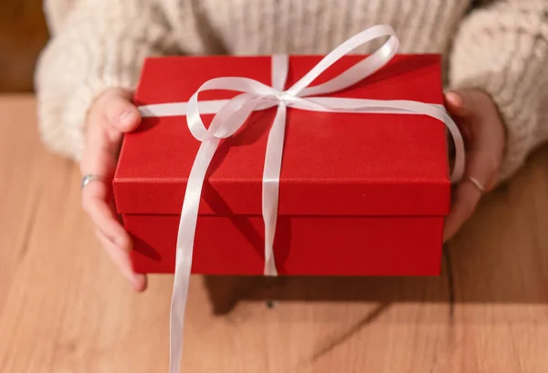 Red gift box with white ribbon in female hands on a wooden table close-up — Stock Photo, Image