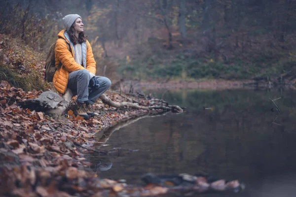 Mujer joven viajera con una chaqueta amarilla y sombrero sentado en un tronco junto al estanque en el bosque nublado de otoño —  Fotos de Stock