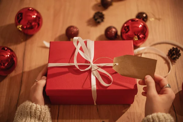 Red gift box with a white ribbon and a wooden tag in female hands on wooden table with festive christmas balls, top view shot — Stock Photo, Image