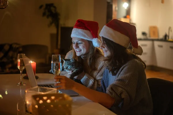 Deux jeunes femmes heureuses et belles copines couple lesbien avec des verres de champagne en chapeaux de Père Noël rouge célèbrent la nouvelle année et Noël à la maison en ligne avec chat et bougies — Photo