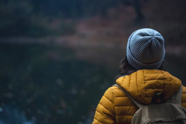 Jonge vrouw reiziger in een gele jas en hoed staan met zijn rug in een mistige herfst bos. — Stockfoto