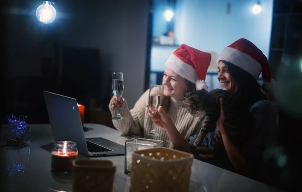 Deux jeunes femmes heureuses et belles copines couple lesbien avec des verres de champagne en chapeaux de Père Noël rouge célèbrent la nouvelle année et Noël à la maison en ligne avec chat et bougies — Photo