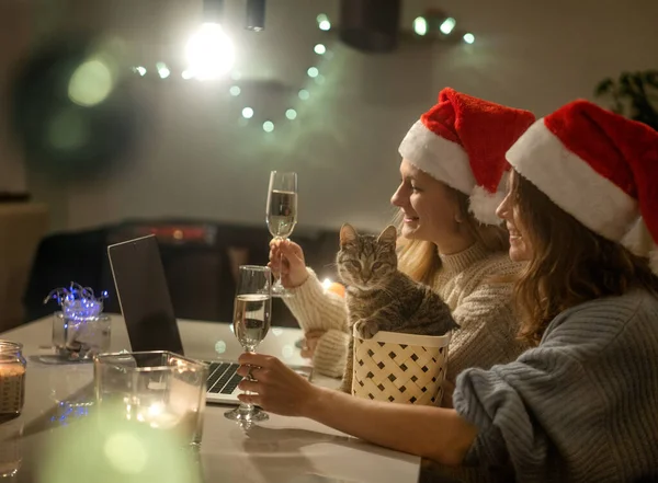 Deux jeunes femmes heureuses et belles copines couple lesbien avec des verres de champagne en chapeaux de Père Noël rouge célèbrent la nouvelle année et Noël à la maison en ligne avec chat et bougies — Photo