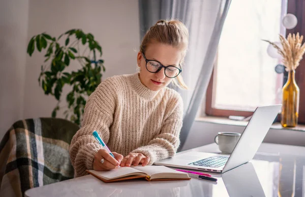 美しい若い幸せな女の子の学生とガラスと暖かい銀はノートパソコンの書き込みで自宅でオンラインで勉強 — ストック写真