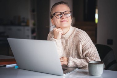 Gözlük takan mutlu genç bir kadın evde oturup dizüstü bilgisayar, internet işi ve eğitim kullanıyor.