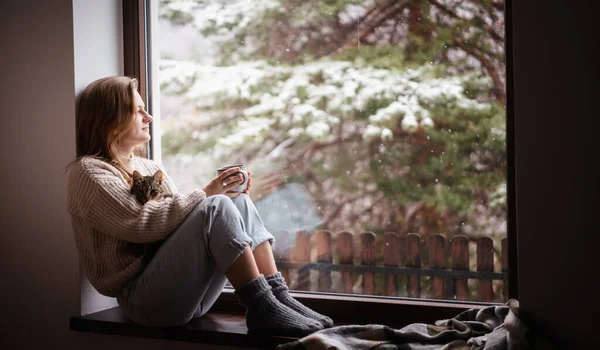 Young cheerful girl sitting at home on the windowsill in a warm sweater drinking coffee with gray kitten in her arms — Stock Photo, Image