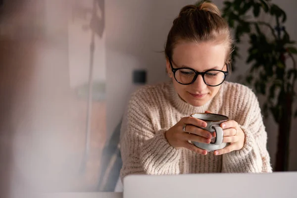 Beautiful young girl blonde woman wearing glasses drinking coffee and looking at the laptop screen — стоковое фото