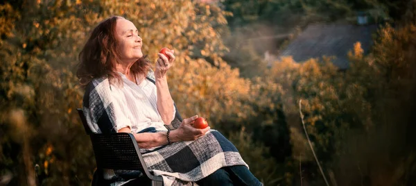 陽気幸せな成熟したシニア女性保持赤リンゴでカントリーハウスガーデンに座って — ストック写真