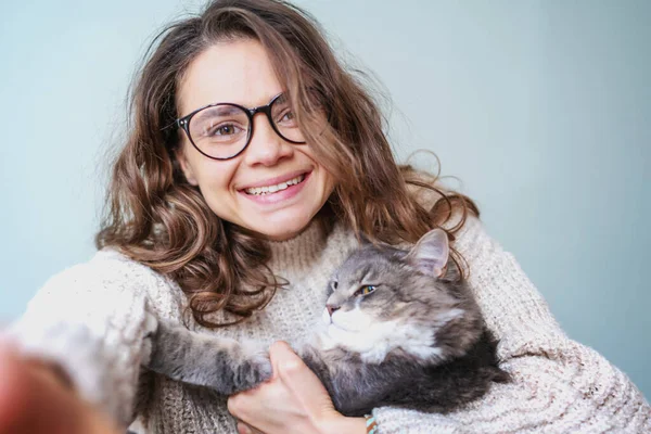 Bonita alegre encaracolado jovem mulher em óculos tomando uma selfie com um gato cinza macio usando smartphone — Fotografia de Stock