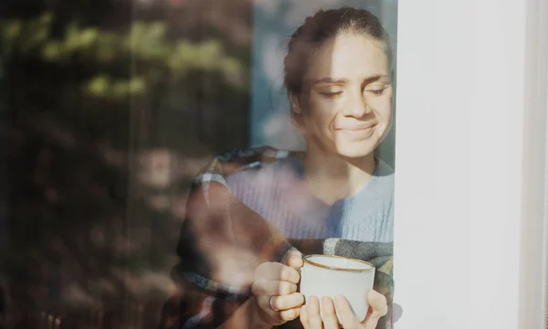 Красивая молодая женщина пьет утренний чай или кофе, сидя у окна в своем загородном доме, в стекле, отражающем сад — стоковое фото