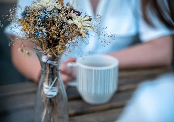 玻璃瓶，花朵干枯，花束美丽。在后面，一个女人在杯子里喝茶 — 图库照片