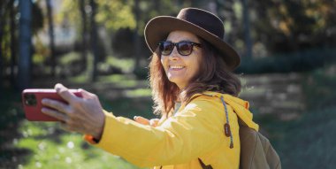 Parkta elinde akıllı bir telefonla şapka ve güneş gözlüğü takan güzel, neşeli, yaşlı bir kadın.