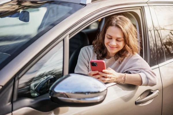 Cheerful young woman sitting in a car in the drivers seat looking into a smartphone, paying for parking