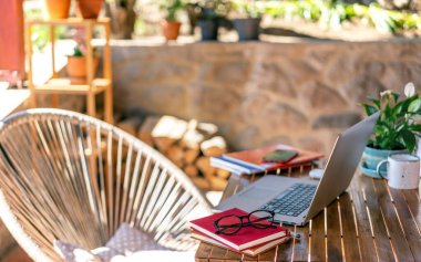 Open laptop on a wooden table, next to glasses on a stack of books on the terrace of a country house on a summer day, remote work and education concept