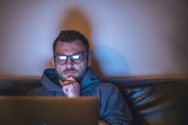 Young attractive serious man in glasses sitting at home at night on an armchair looking into a laptop. Workaholic insomnia hacker concept