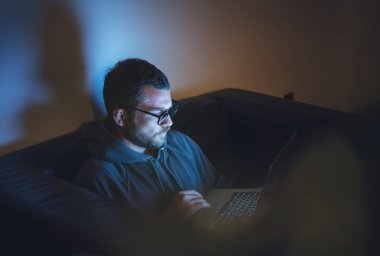 Young attractive serious man in glasses sitting at home at night on an armchair looking into a laptop. Workaholic insomnia hacker concept