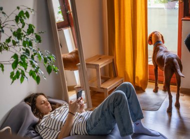 Young woman lying on bag chair at home using smartphone, dog standing at the door waiting for a walk