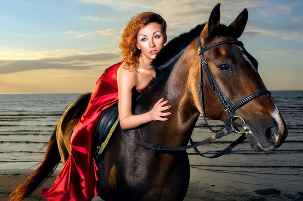 Γυναίκα σε ένα κομψό κόκκινο φόρεμα με ένα άλογο στην παραλία — Φωτογραφία Αρχείου