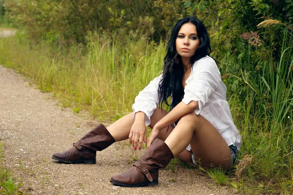 Sexy girl in denim shorts and white shirt sitting by the countryside road — Stock Photo, Image