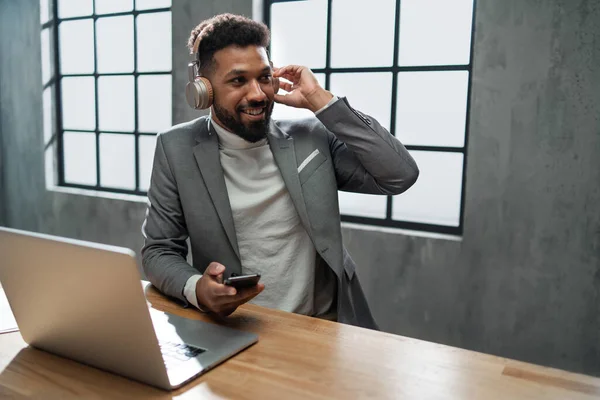 Gelukkig jong Afrikaans Amerikaans zakenman met smartphone en headset werken op een laptop binnen in kantoor — Stockfoto