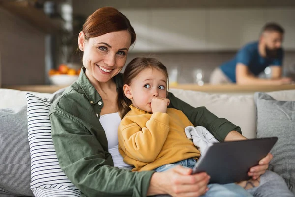 Madre con hija pequeña usando tableta en el interior de casa, mirando a la cámara, la vida cotidiana y la oficina en el hogar con concepto de niño. — Foto de Stock