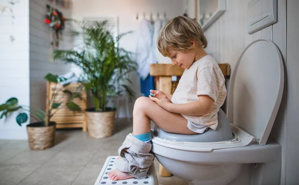 Tampilan samping anak kecil yang lucu duduk di dalam toilet di rumah, menggunakan smartphone. — Stok Foto