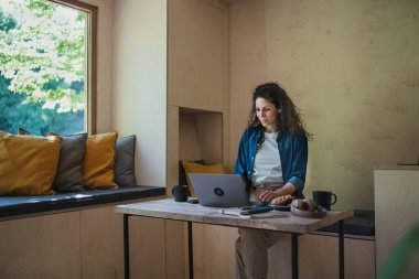 Kahveli mutlu genç bir kadın evde dizüstü bilgisayarla oturuyor ve çalışıyor, hafta sonu uzakta ve bireysel seyahat konsepti.