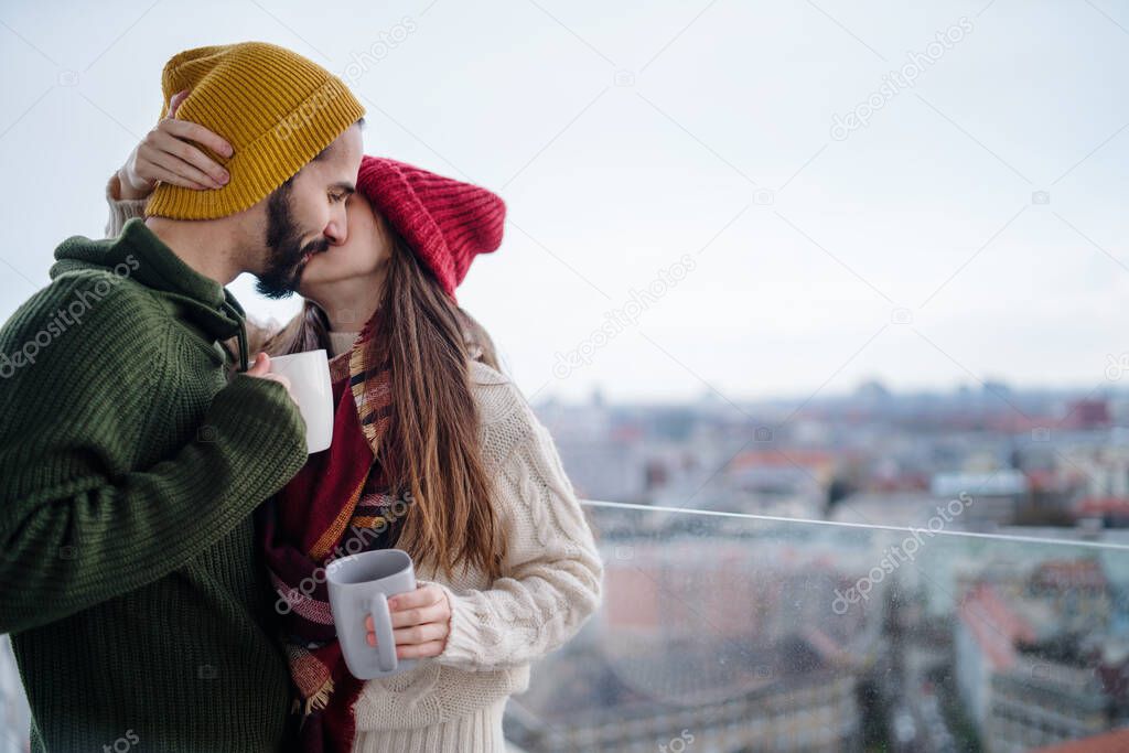 Young couple with coffee standing and kissing outdoors on balcony with urban view