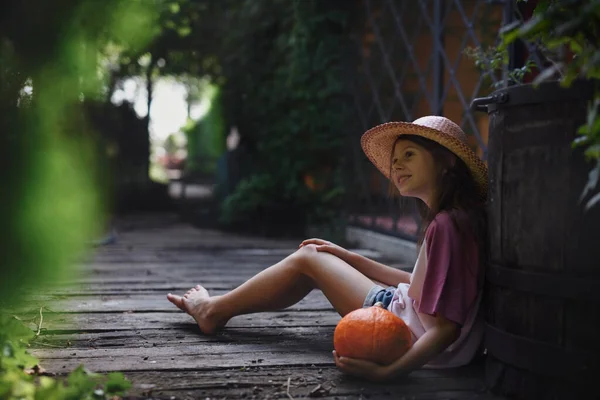 Niña en sombrero sentada y sosteniendo calabaza orgánica al aire libre en la granja. —  Fotos de Stock