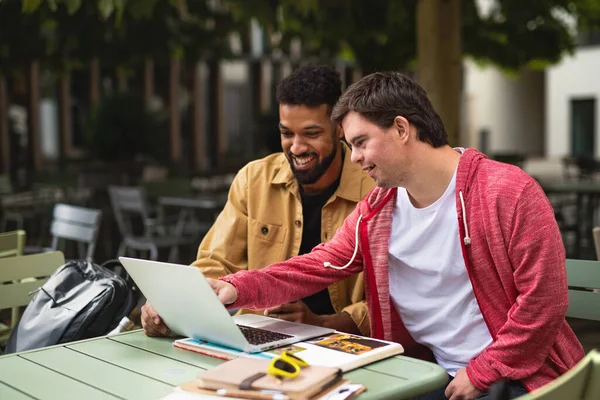 Mladý muž s Downovým syndromem se svým mentorským přítelem sedí venku v kavárně pomocí notebooku. — Stock fotografie