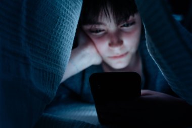 Teen girl using smartphone, hiding under blanket at nigh, social networks cocnept. clipart