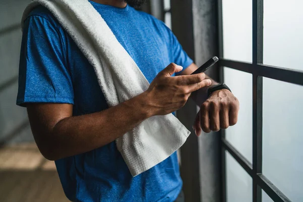 Oigenkännlig ung afroamerikansk idrottsman står på gym, med hjälp av smartphone och smartwatch — Stockfoto