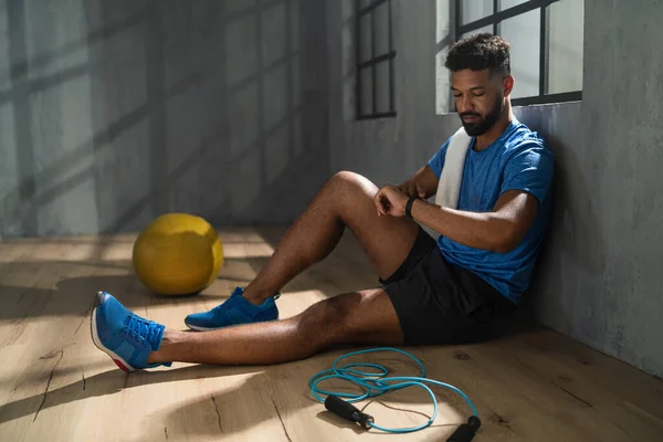 Молодой афроамериканский спортсмен сидит и использует умные часы в спортзале, тренировки — стоковое фото
