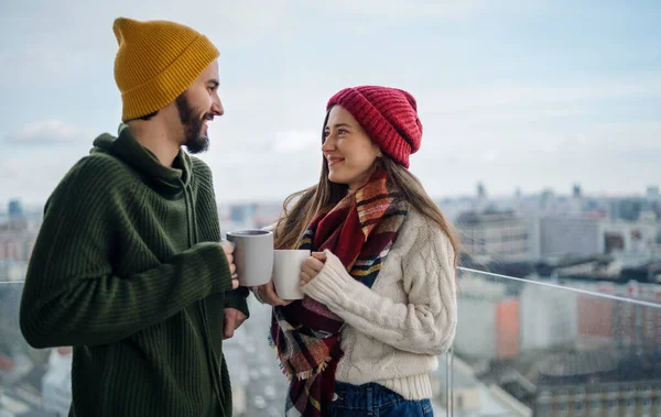 一对年轻的情侣，站着喝咖啡，彼此凝视着外面的阳台，欣赏都市美景. — 图库照片