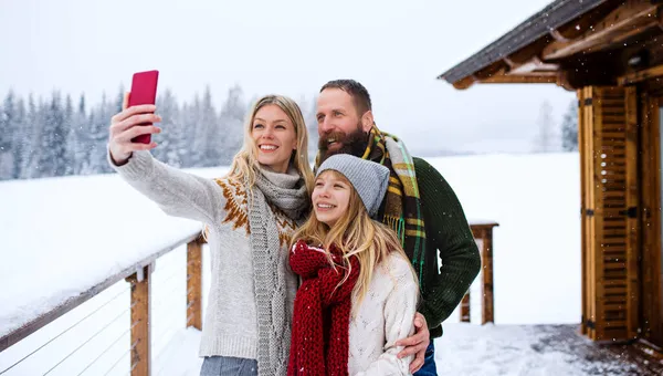 Lycklig familj med liten dotter som tar selfie vid fjällstugan utomhus på vintern. — Stockfoto