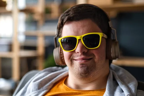 Primo piano di giovane felice con sindrome di Down con occhiali da sole e cuffie seduti al chiuso — Foto Stock